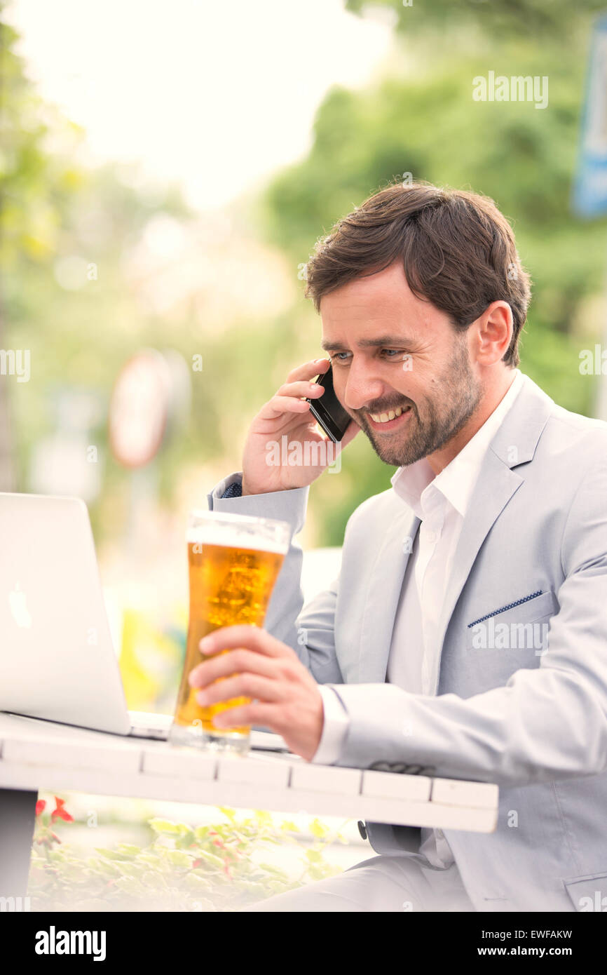 Happy businessman using mobile phone and laptop while holding beer glass at outdoor restaurant Stock Photo