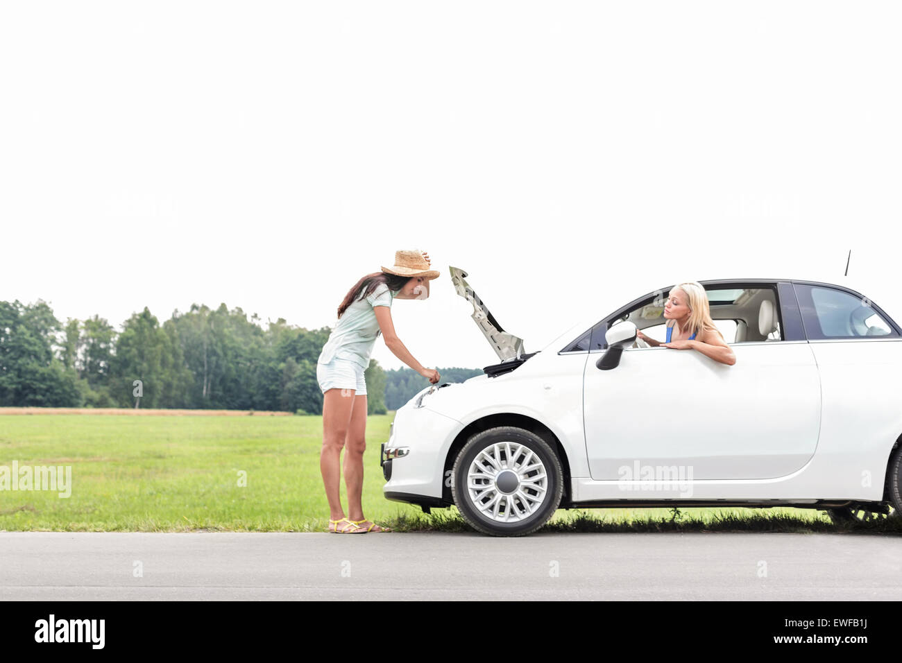 Woman looking at friend repairing broken down car on country road Stock Photo