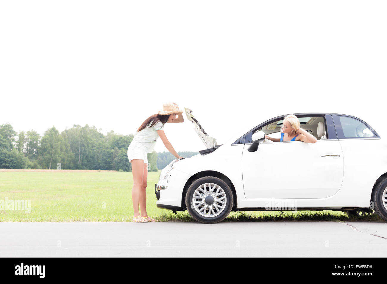Woman looking at female friend repairing broken down car on country road Stock Photo