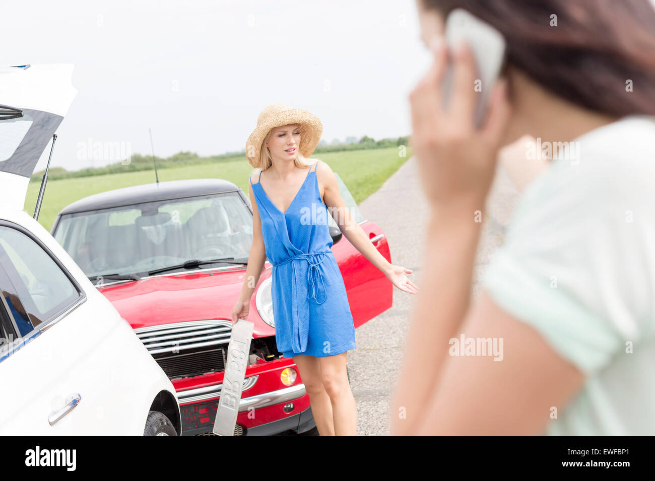 Angry woman standing by damaged cars with female using cell phone in foreground Stock Photo