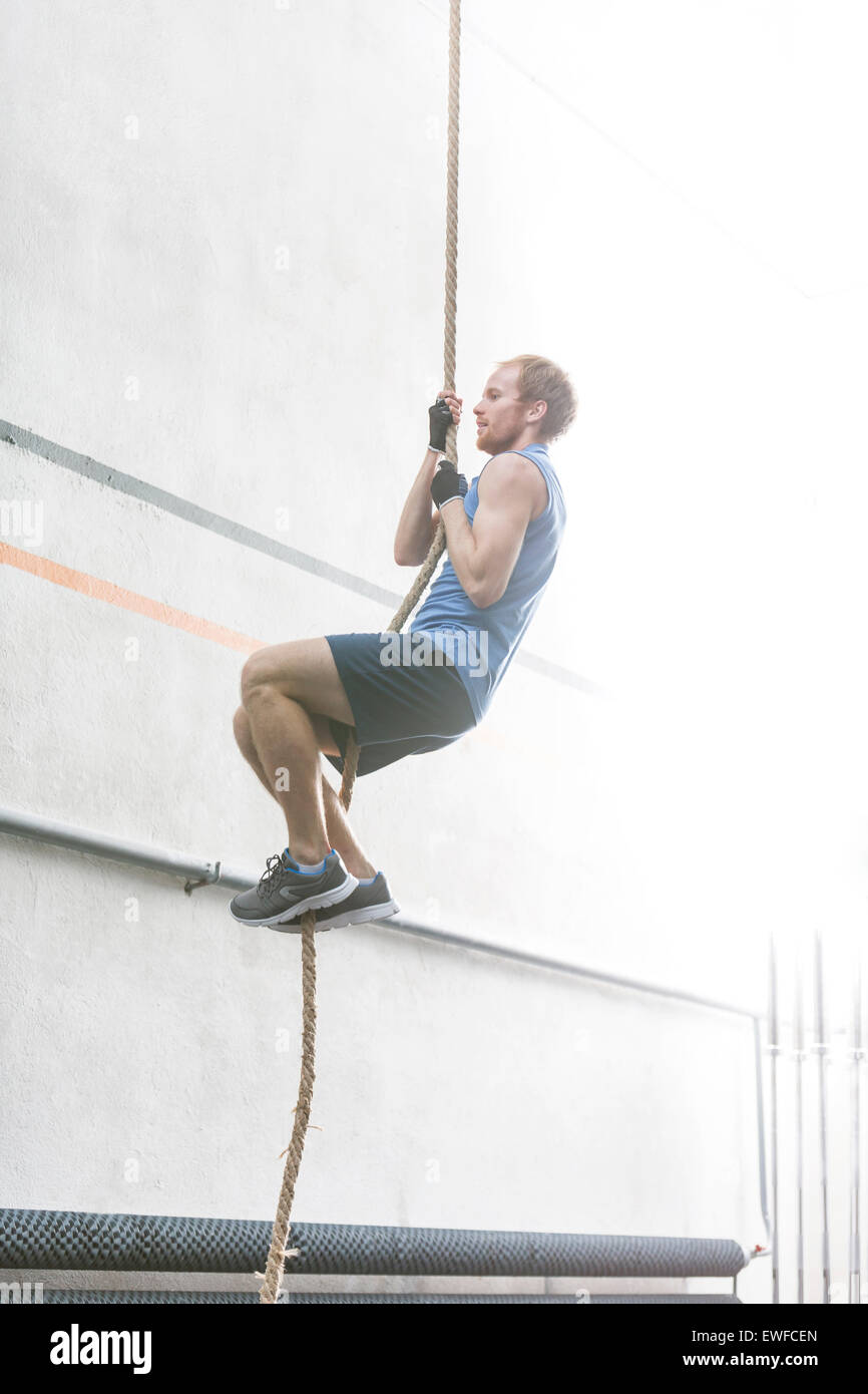 Side view of determined man climbing rope in crossfit gym Stock Photo