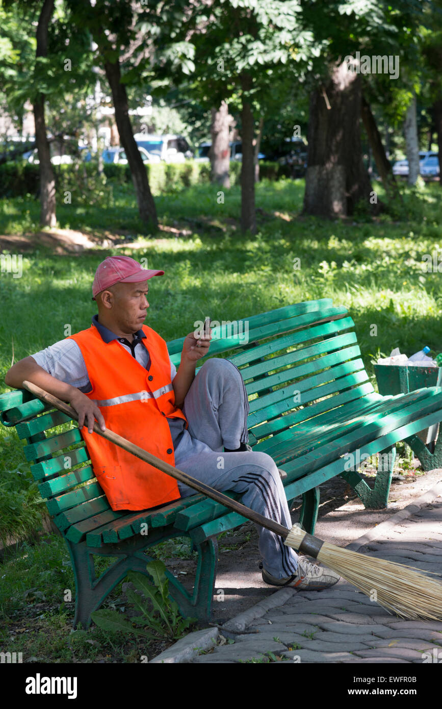 Street sweeper on a bench with his smartphone. Public garden. Bishkek. Kyrgyzstan. Central Asia. Stock Photo