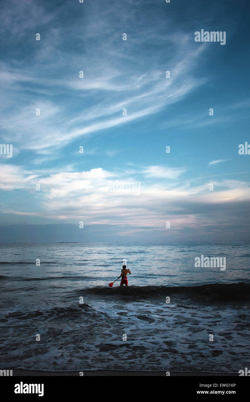A man stands in the ocean, with the blue sky and clouds reflected on ...