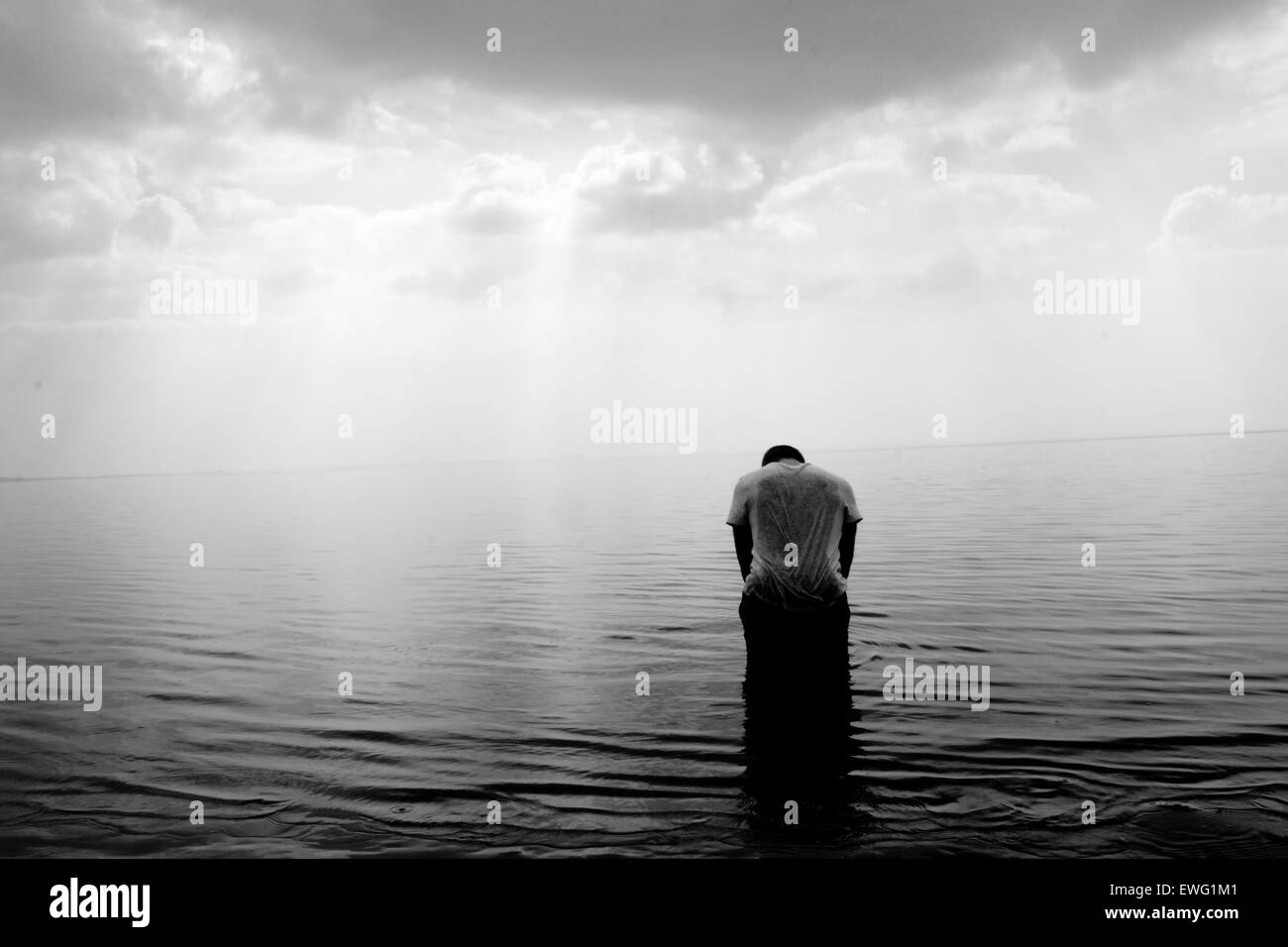 man bowing his head in an ocean with sun coming through the sky Stock Photo