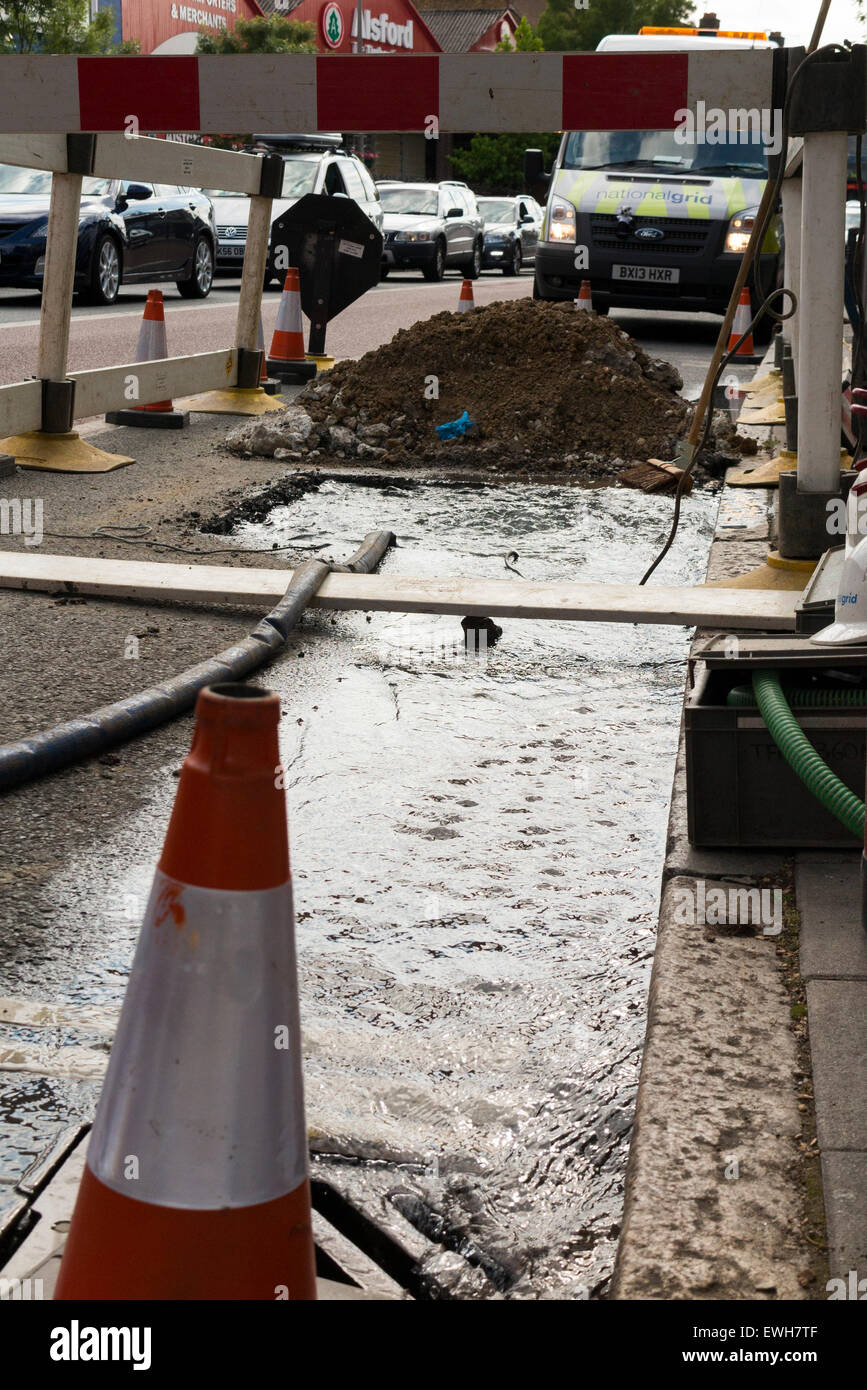 Broken / burst water main leaking pipe allows water to leak and emerge through the street & flow in the in gutter / road. UK Stock Photo