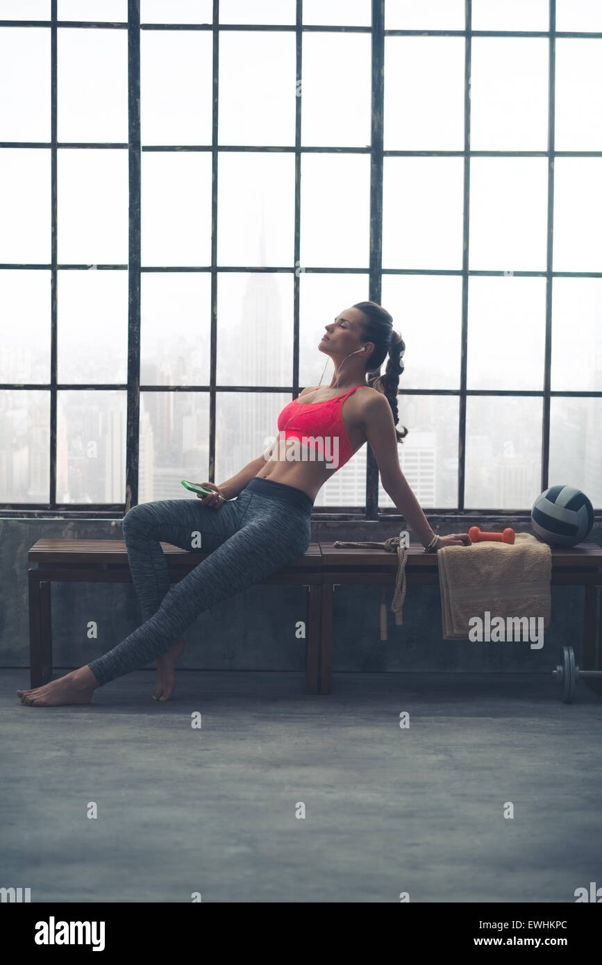 A woman is relaxing on a bench by the window in a big loft gym. Lost in music, she is stretching herself out in a relaxed way, leaning back, head tilted upwards. Stock Photo