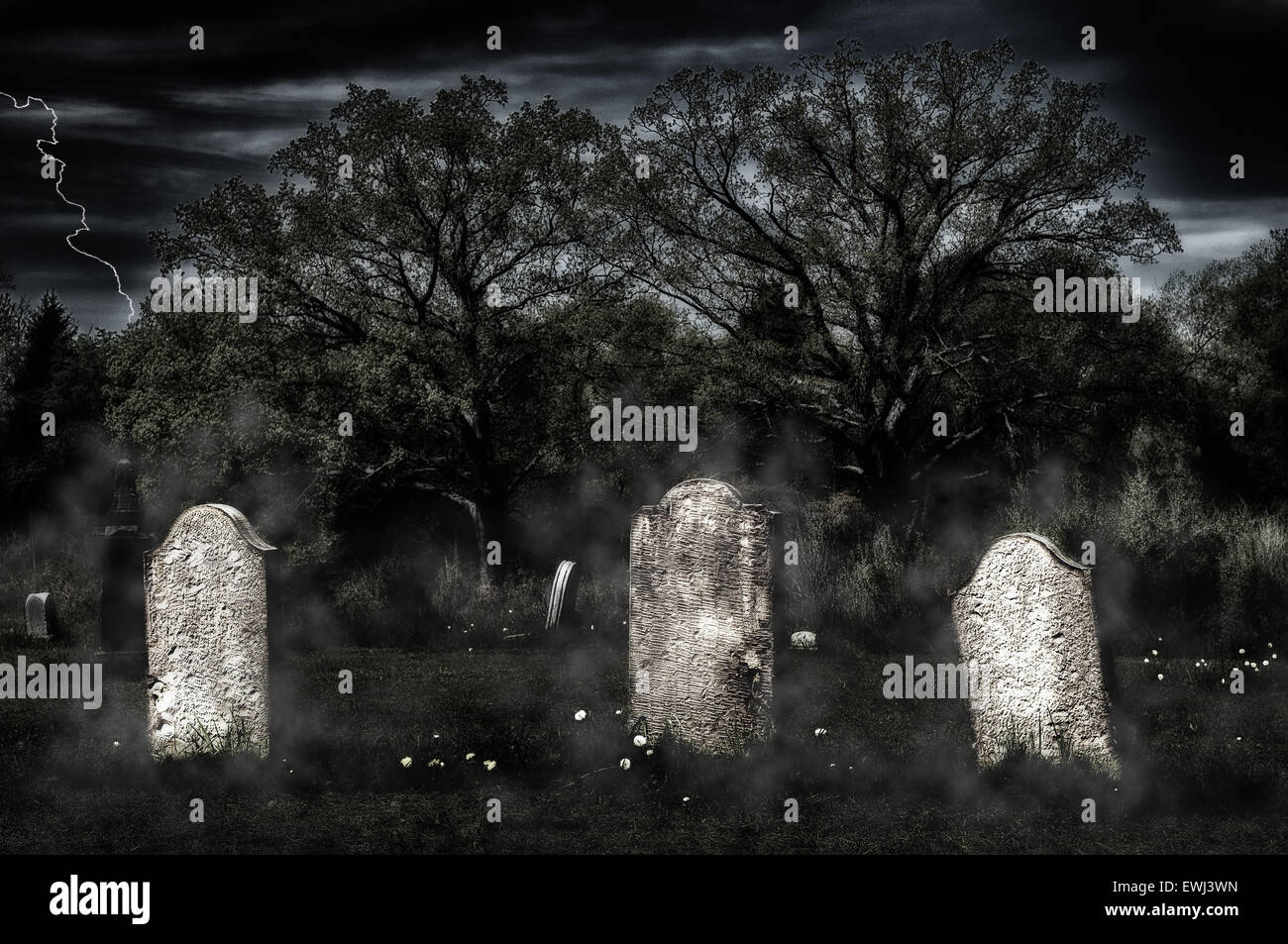 Weathered headstones in an old cemetery glow in an eerie light in the gloom, with a grainy sky and fog wisps Stock Photo