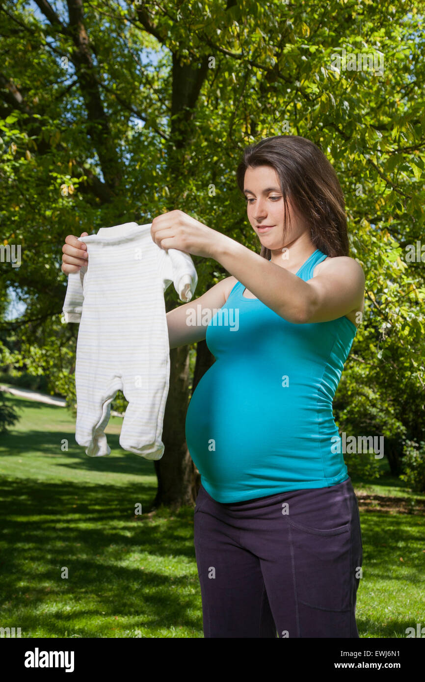 pregnant young woman blue shirt with white baby clothes on her hands and green trees background Stock Photo
