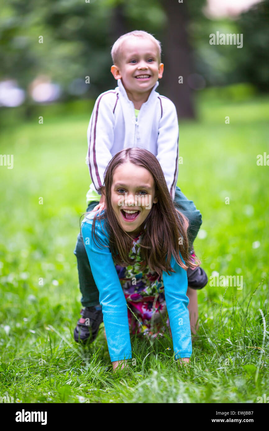 Girl with her little brother playing in the green grass. Little boy riding on back a sister. Stock Photo