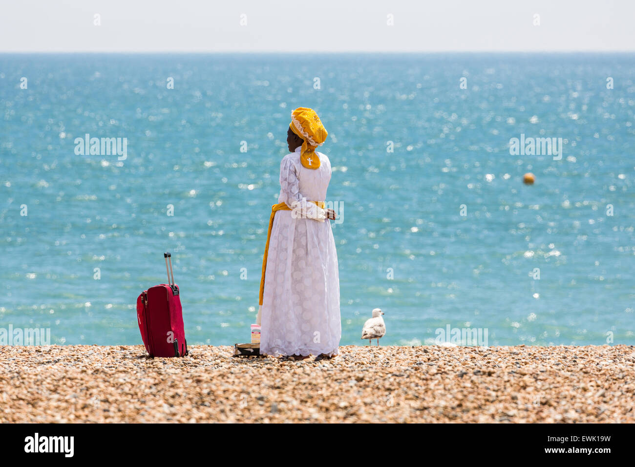 Man in unusual white dress and yellow headwear, with suitcase on the beach in Brighton, East Sussex, UK Stock Photo