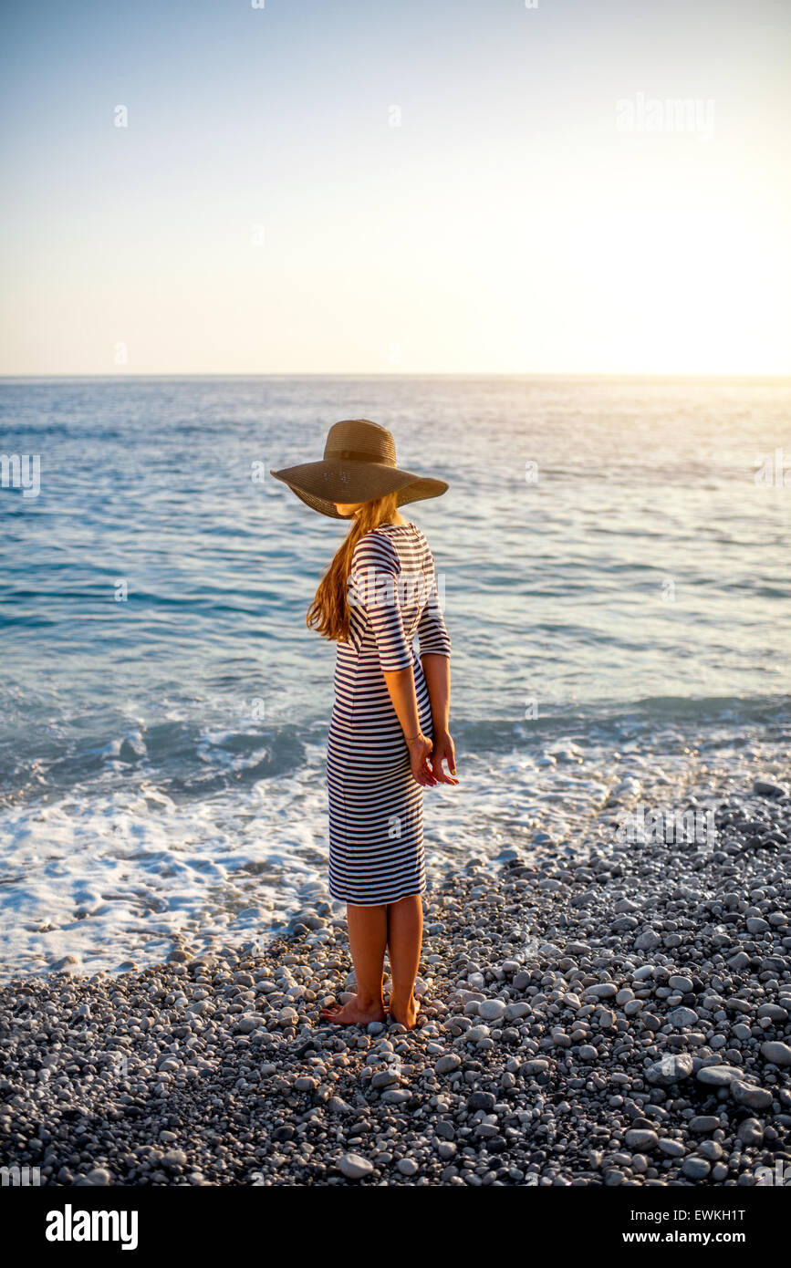 Young and elegant woman in stripped dress with a hat standing on the beach on sunset. Back view with copy space Stock Photo