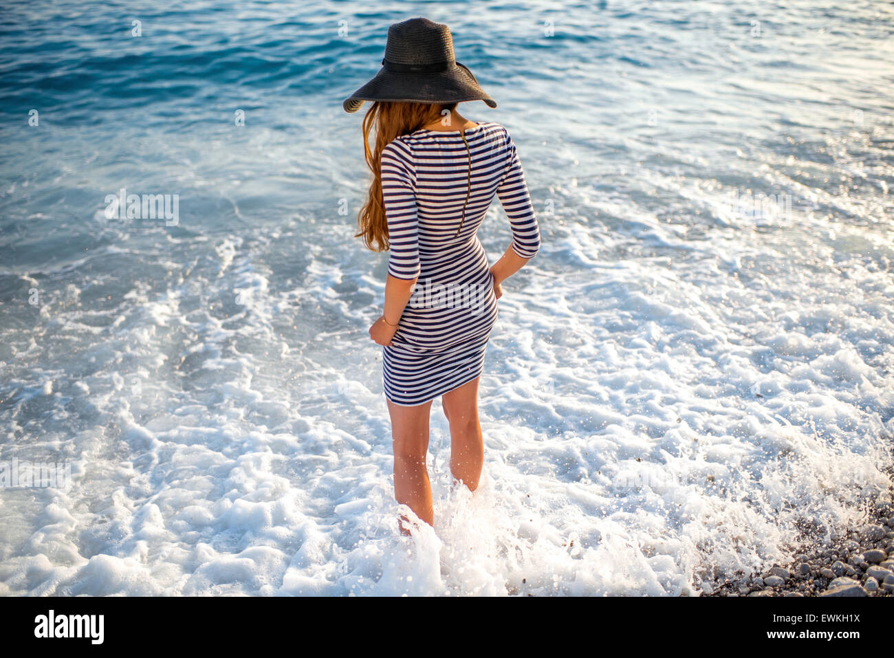 Young and elegant woman in stripped dress with a hat standing in the water with white foam on sunset. Back view Stock Photo