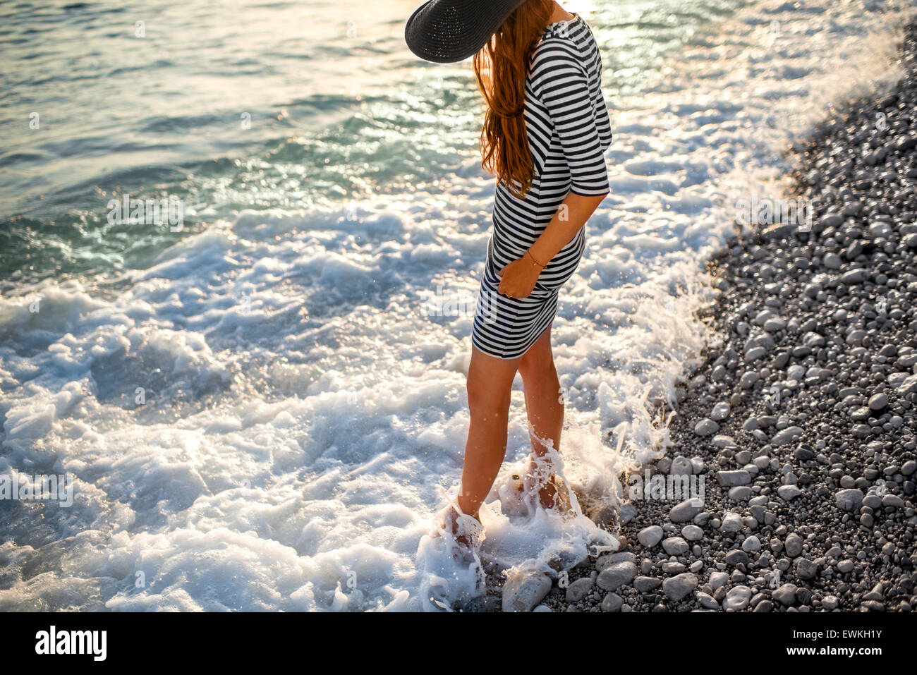 Young and elegant woman in stripped dress with a hat standing in the water with white foam on sunset. Back view Stock Photo
