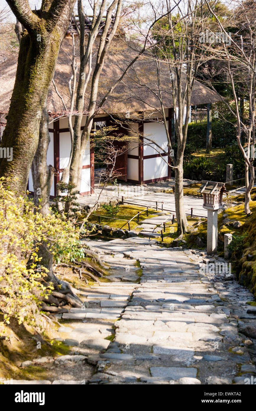 Stone paved footpath leading through embankment with trees on to a small wood and white plaster thatched roof gatehouse at Jojakkoji temple in Kyoto. Stock Photo