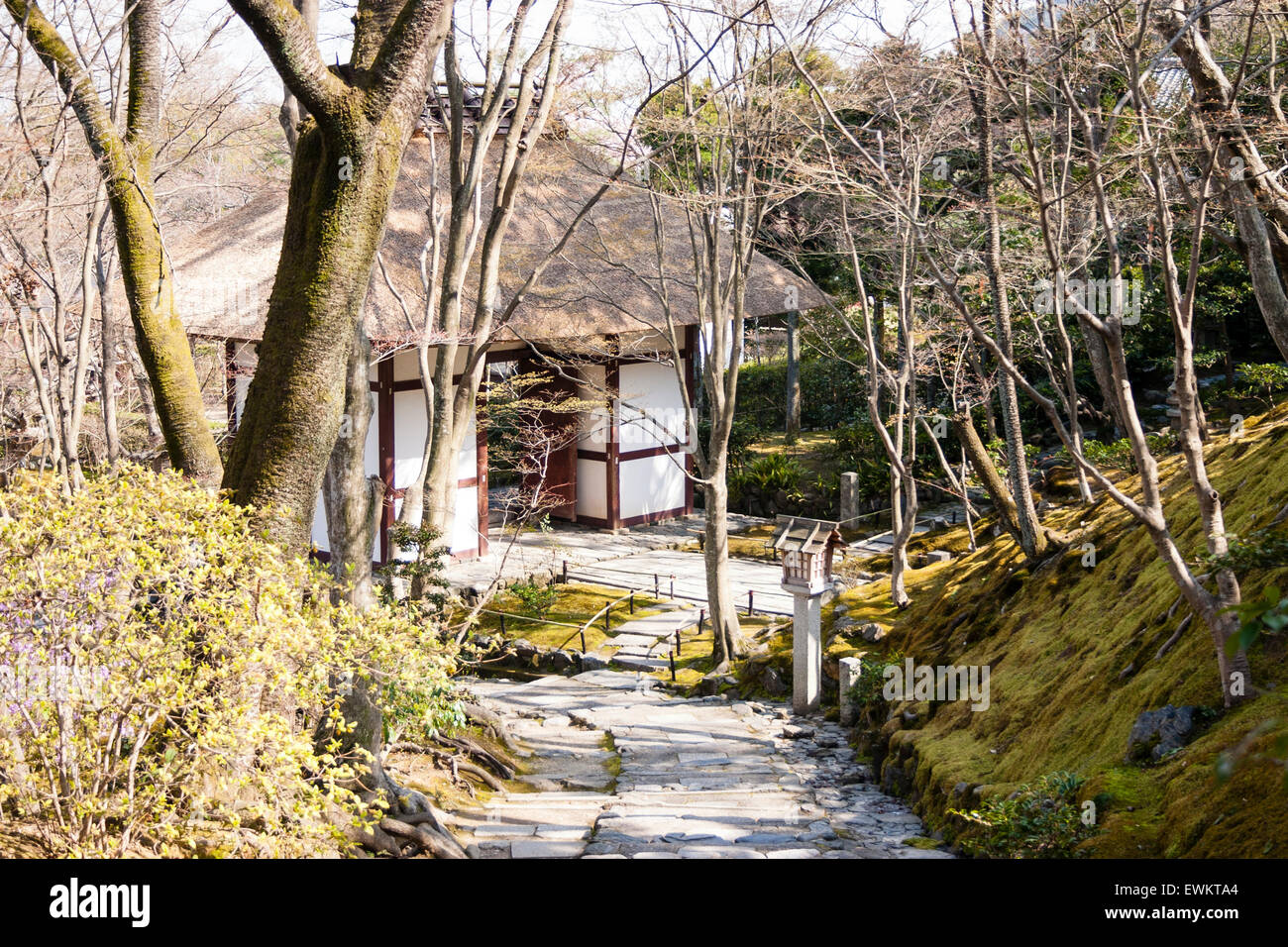 Stone paved footpath leading through embankment with trees on to a small wood and white plaster thatched roof gatehouse at Jojakkoji temple in Kyoto. Stock Photo