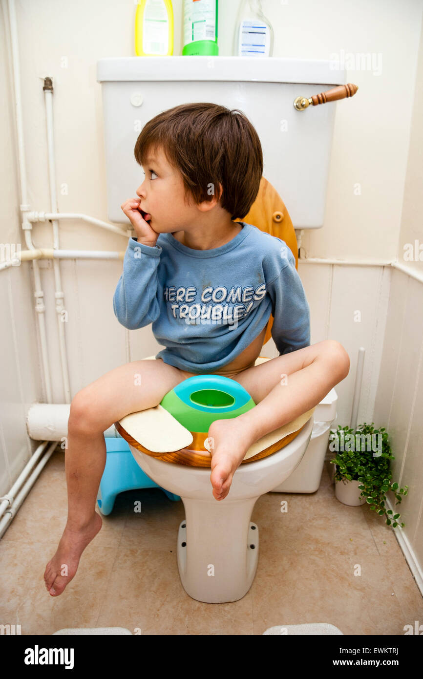 Caucasian young child, boy, 4-5 year old, facing viewer and sitting on baby seat on top of toilet during toilet training. Looking at side wall. Stock Photo