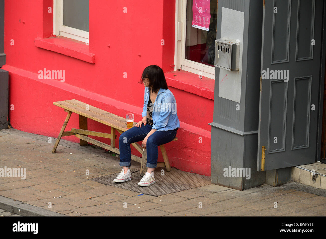 Young woman sitting on bench outside pub with glass of beer, in Londonderry (Derry), Northern Ireland Stock Photo