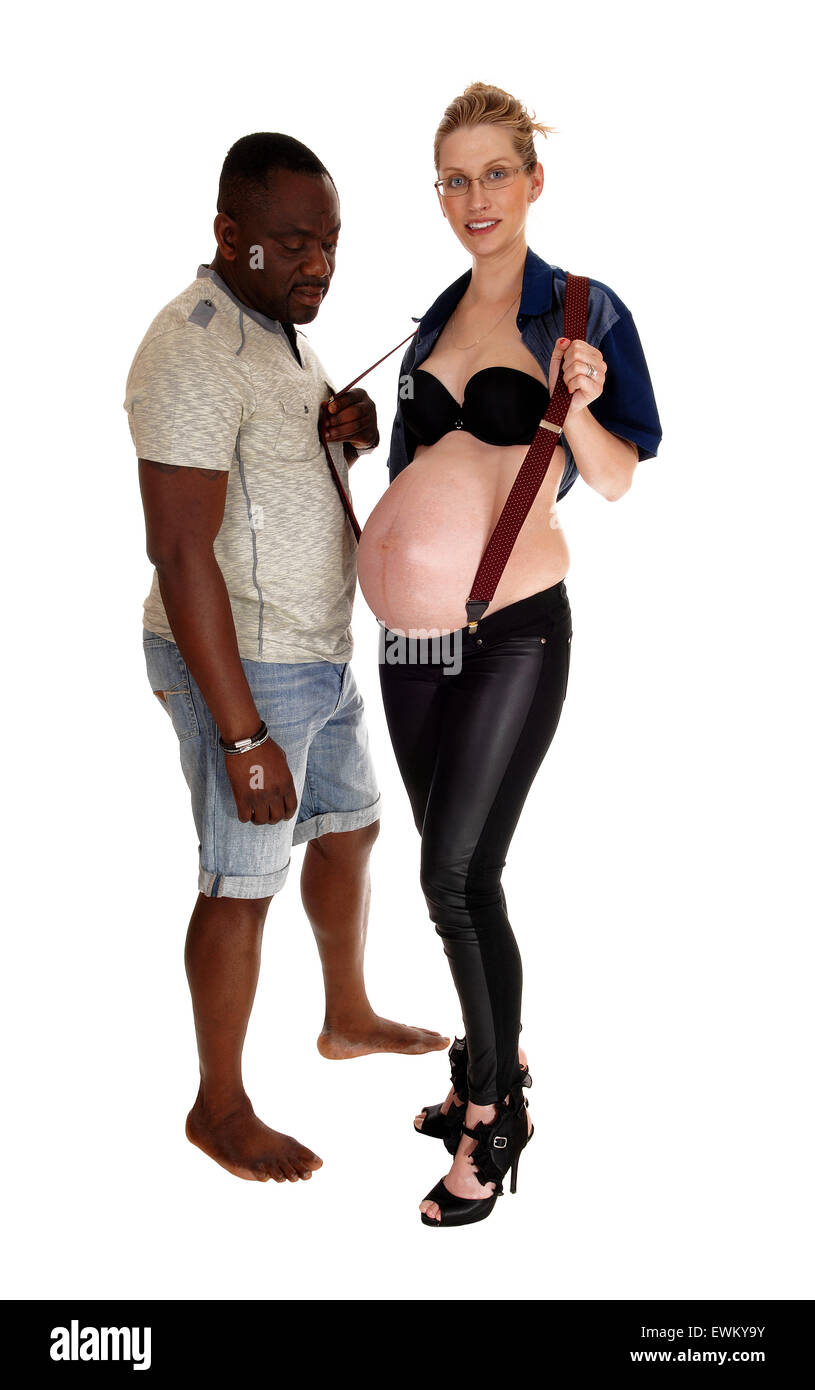 A happy Caucasian pregnant woman with her baby tummy, holding her suspender, her African man is looking on, isolated for white b Stock Photo