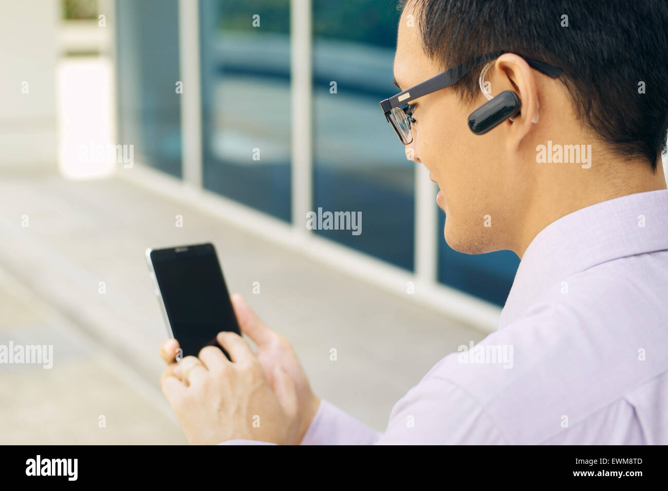 Young chinese businessman typing phone number on smartphone and talking with bluetooth headset device Stock Photo