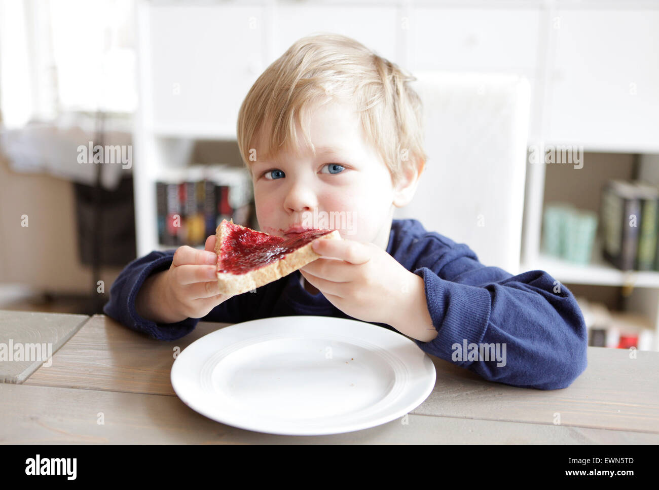 a blond boy sitting at a table and eating a jam sandwich Stock Photo