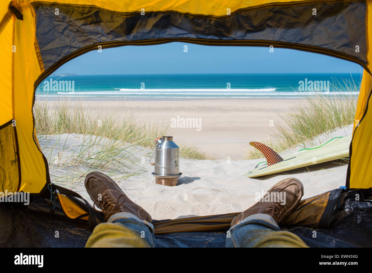 View from a tent on the beach with a surfboard, stove and the sea in the distance. Stock Photo