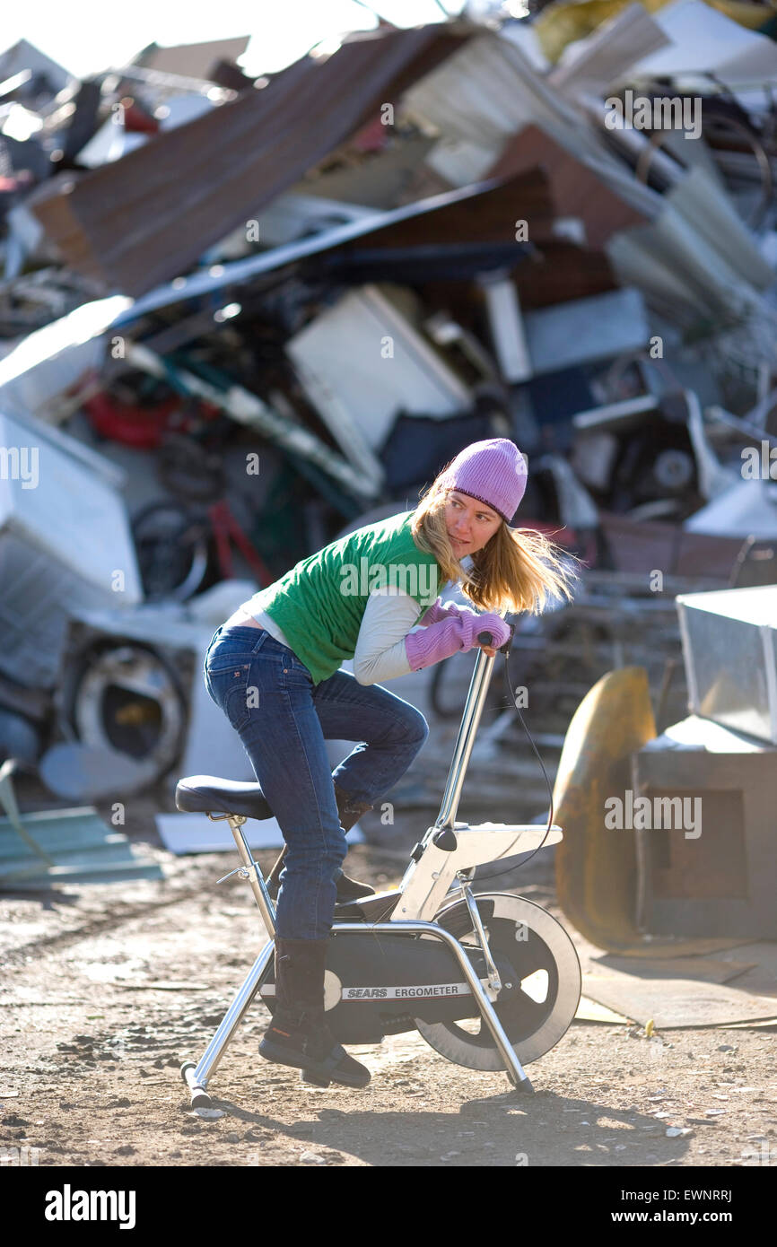 Adult woman riding a junked stationary bike in front of a scrap metal pile Stock Photo