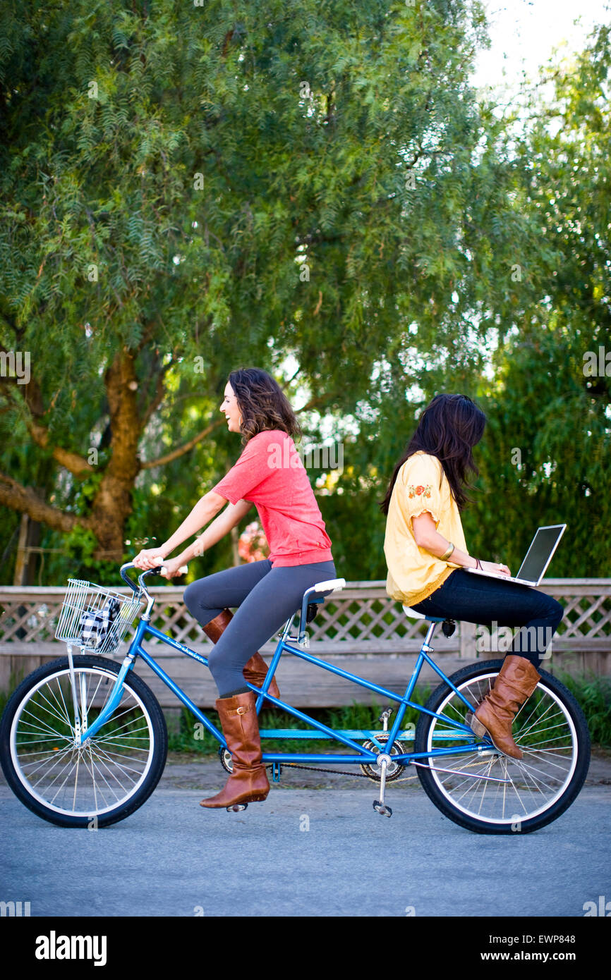 Two women riding a tandem bike while the woman on back surfs the internet on a laptop Stock Photo