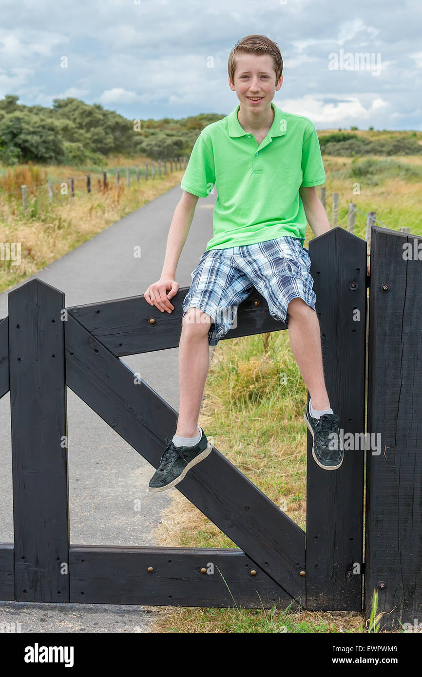 Caucasian teenage boy sitting on black wooden fence in nature as tourist enjoying vacation Stock Photo