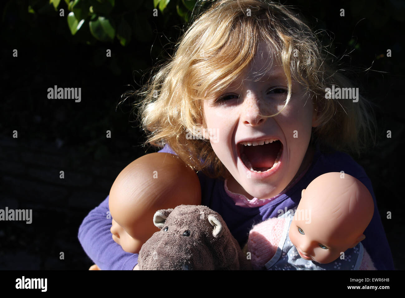 A six year old girl screams with joy as she hugs her most valuable posessions: two dolls and a stuffed hippopotamus Stock Photo