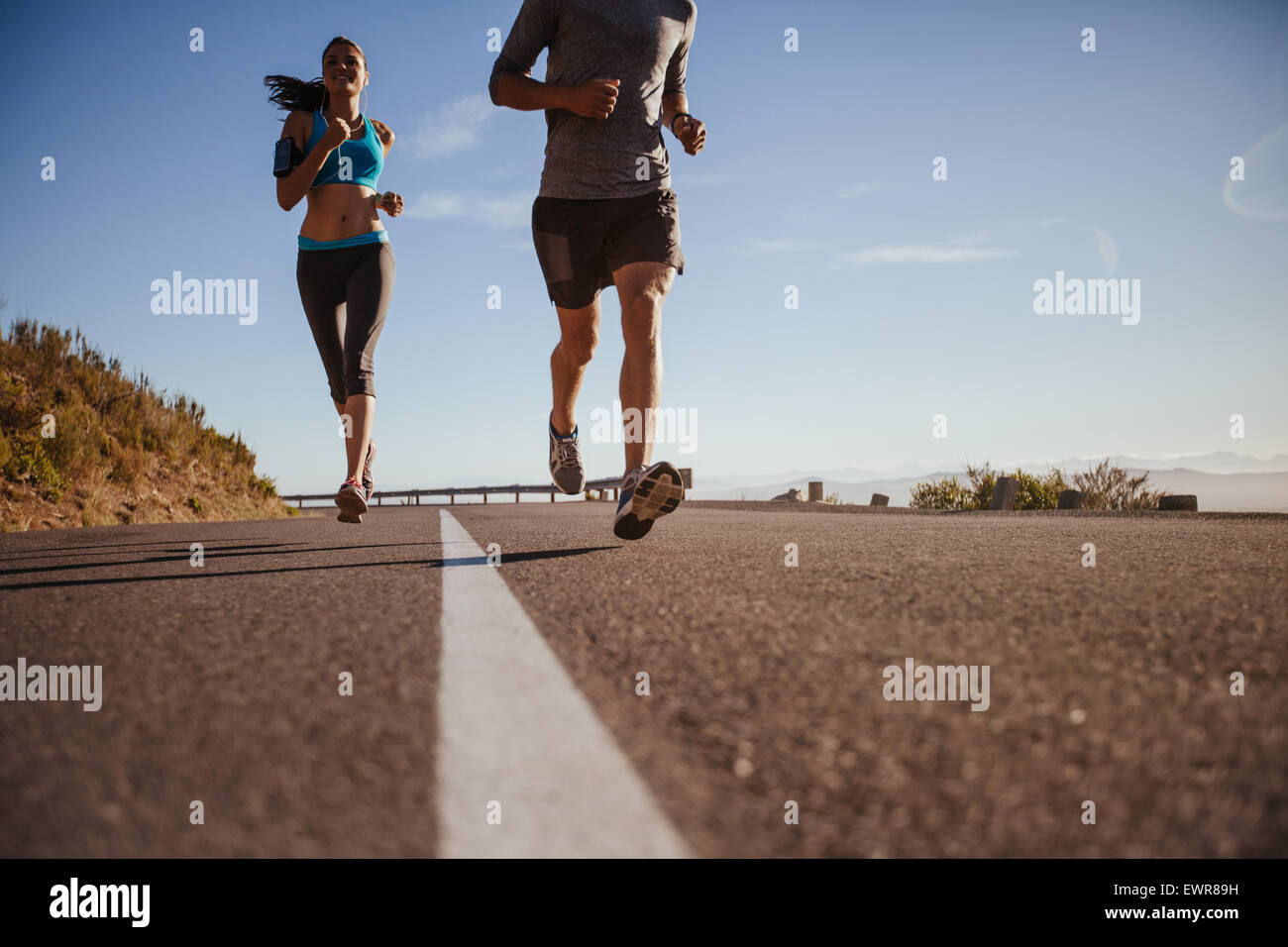 Low angle shot of young woman running on road with man in front on a summer morning. Runners training on country road. Stock Photo