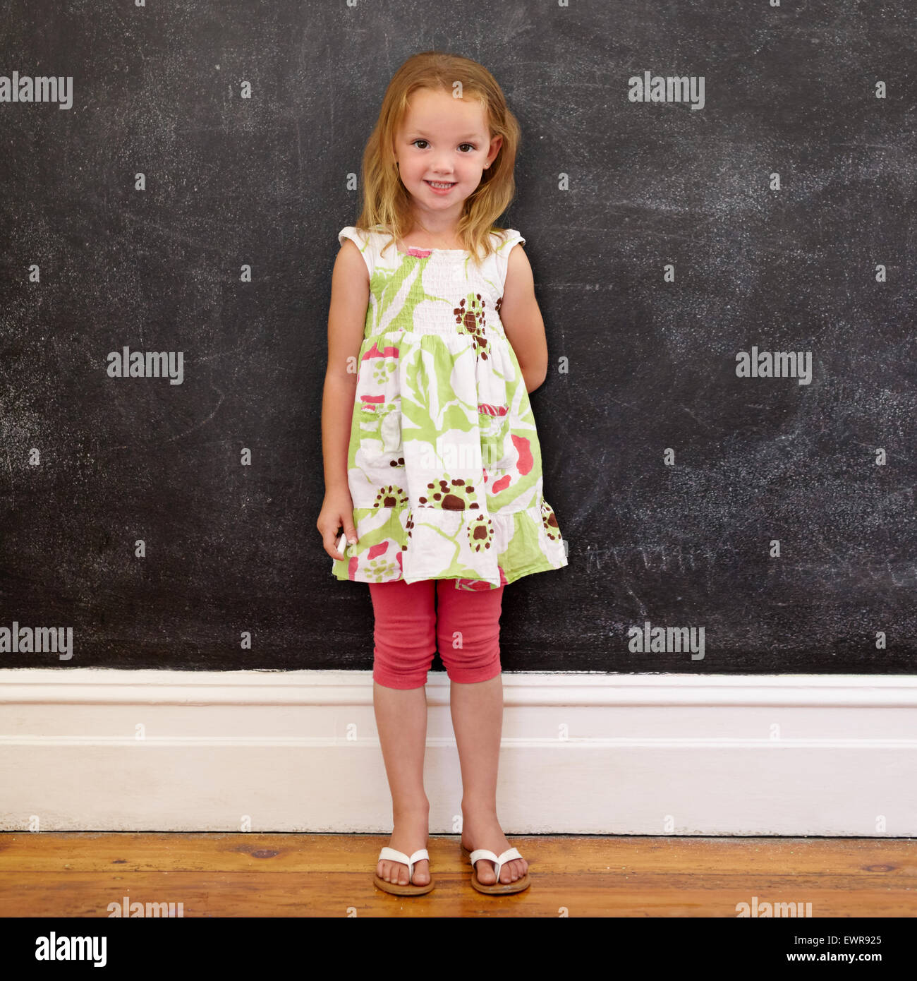 Full length shot of beautiful little girl standing against blackboard at home. She is holding a chalk looking at camera. Stock Photo