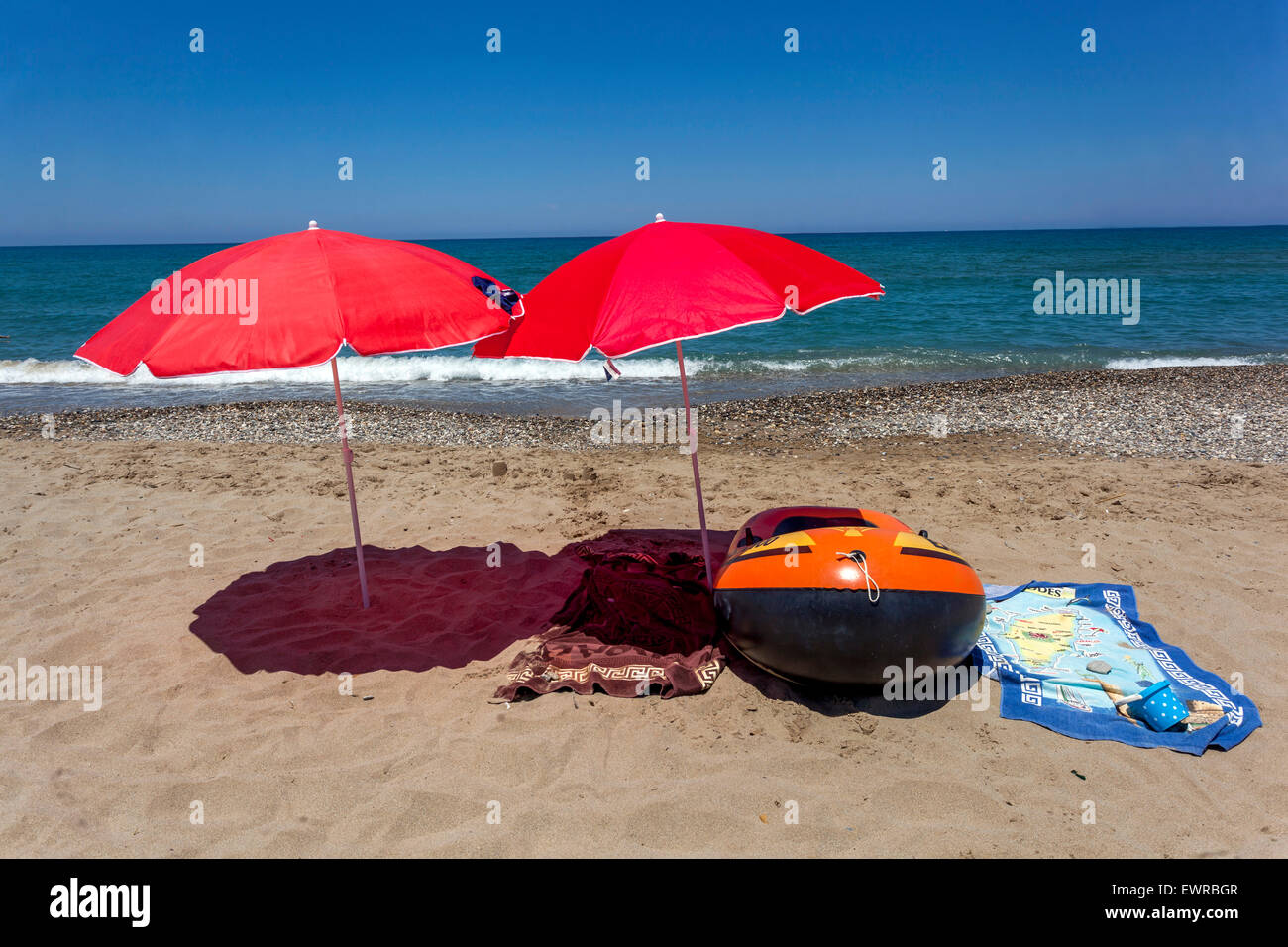 Two red beach umbrellas on a beach of Rethymno, Crete, Greece beach parasols cast a shadow, Still life on the beach Inflatable boat and towels, nobody Stock Photo