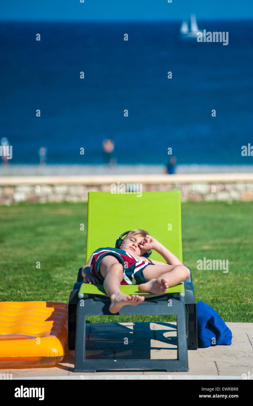A boy lying on a sun lounger listening to music on his mp3 player through  headphones whilst on holiday in the Greek Islands Stock Photo - Alamy