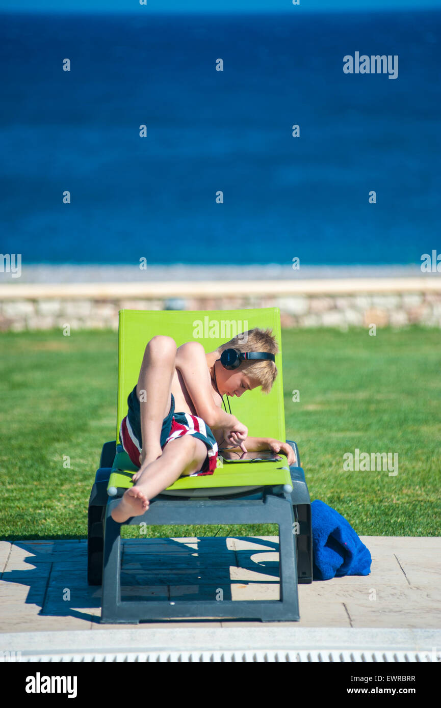 A boy lying on a sun lounger listening to music on his mp3 player through headphones whilst on holiday in the Greek Islands Stock Photo
