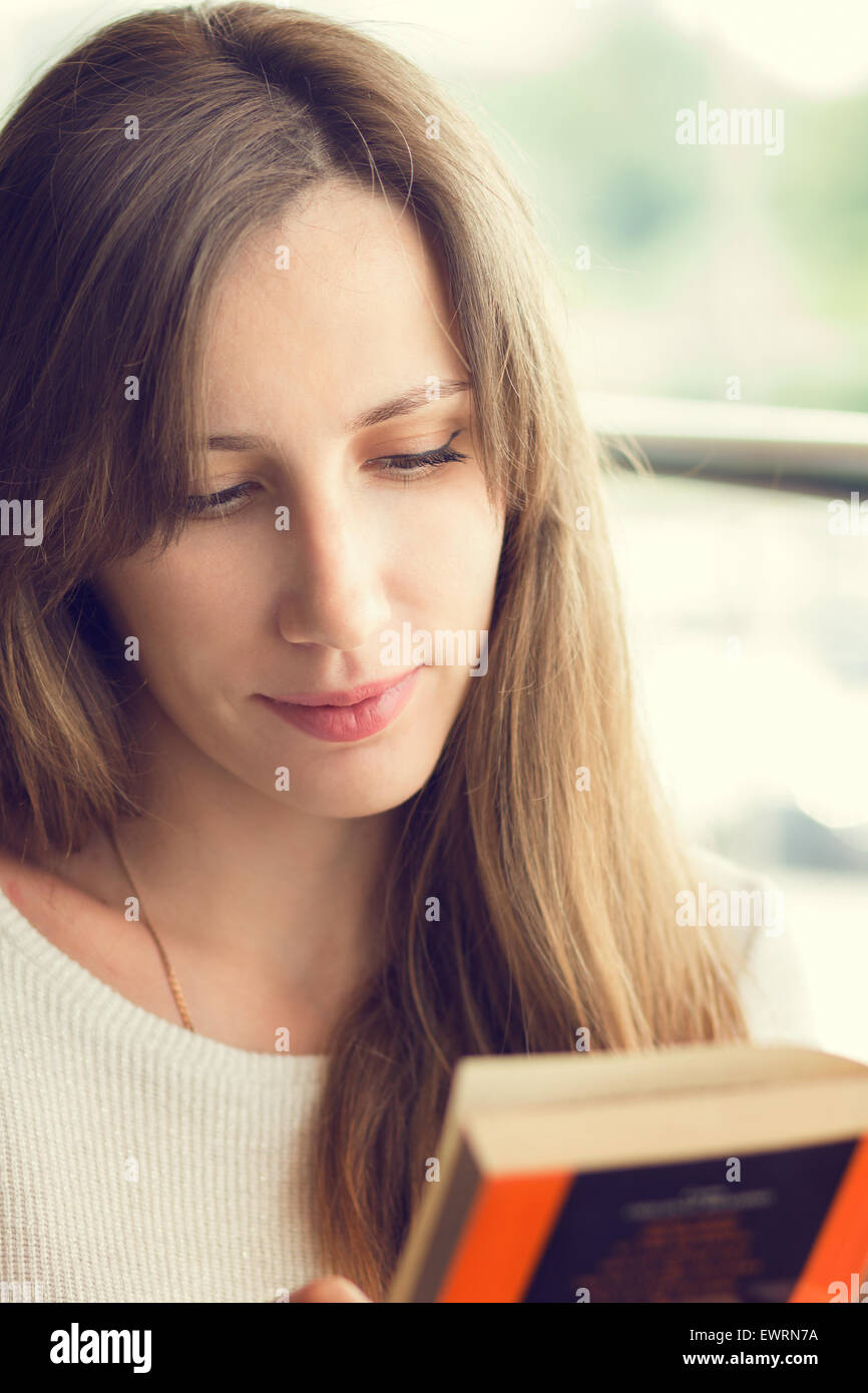 Young beautiful woman reading a book in a cafe near window. Warm color toned image Stock Photo