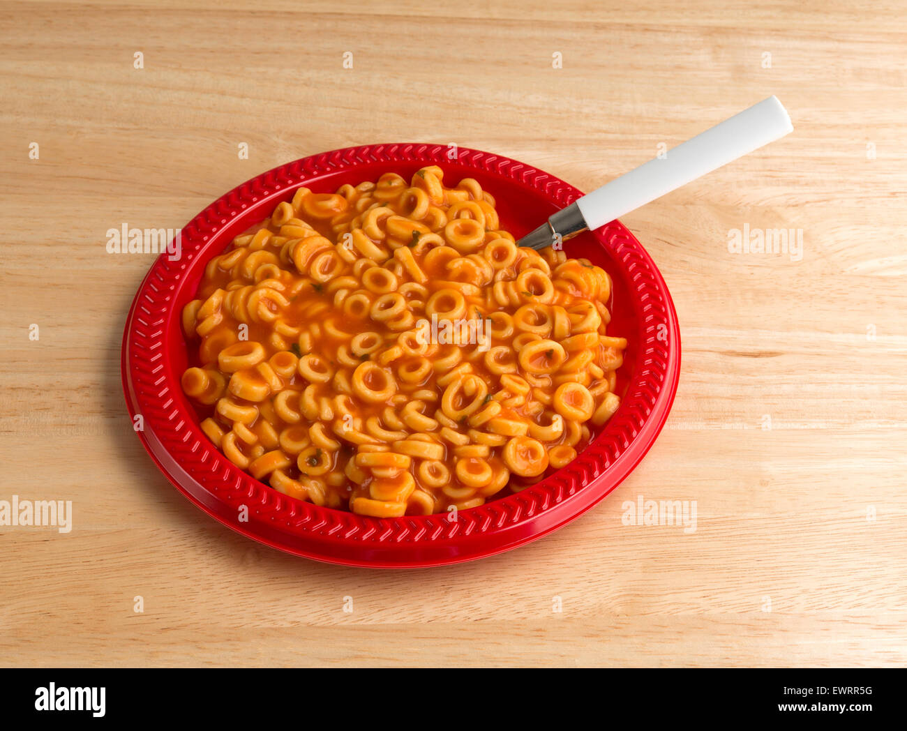 Side view of cooked canned round spaghetti pasta in tomato sauce on a red plate with a spoon illuminated by natural light. Stock Photo
