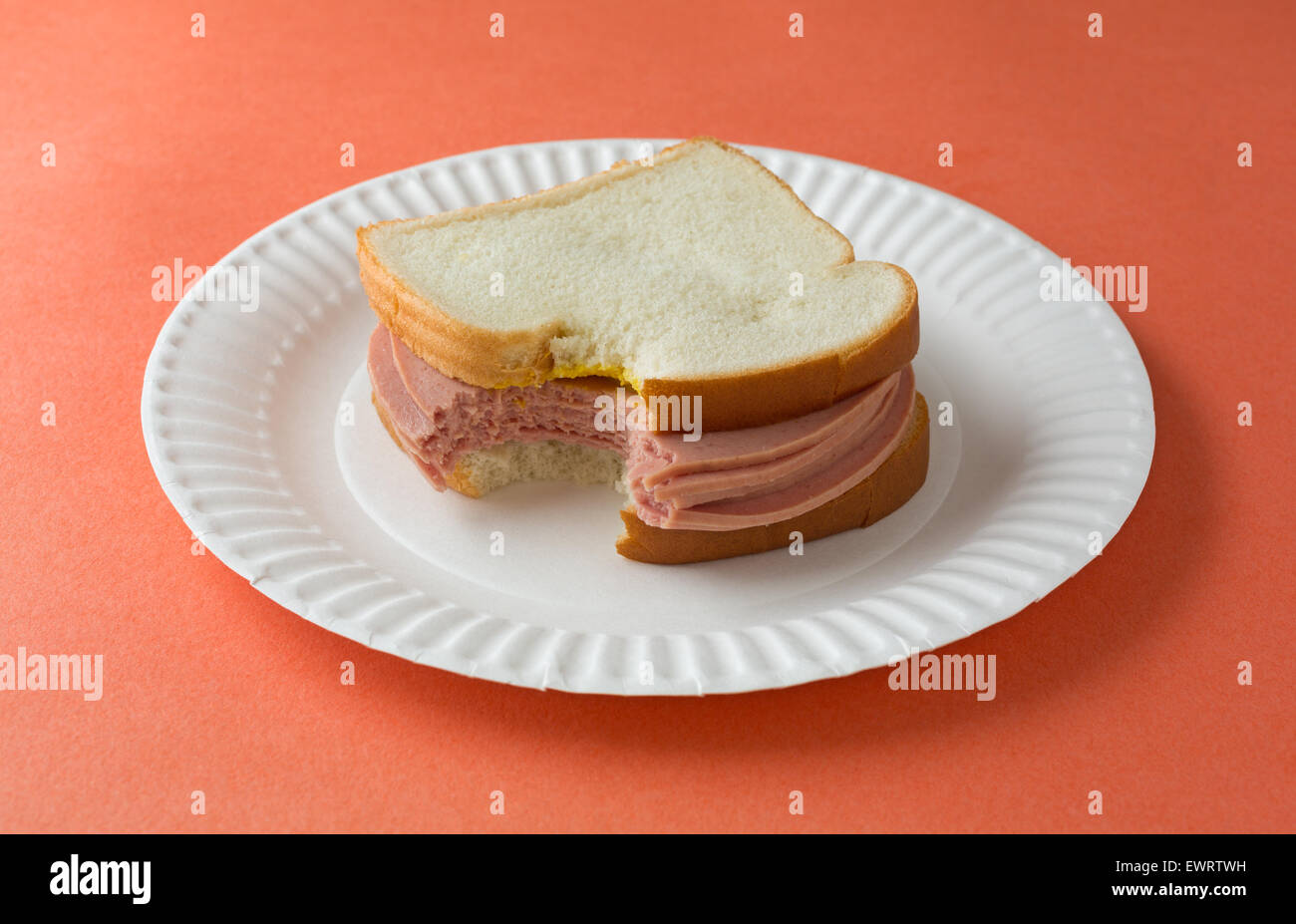 A bitten bologna sandwich with mustard and white bread on a paper plate