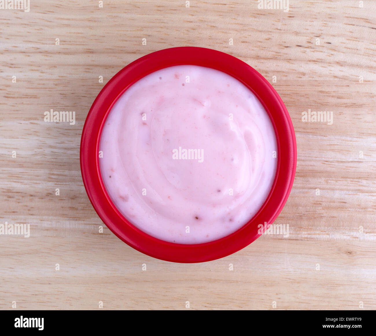 Top view of a small serving of creamy strawberry yogurt in a red bowl on top of a wood table top illuminated by window light. Stock Photo