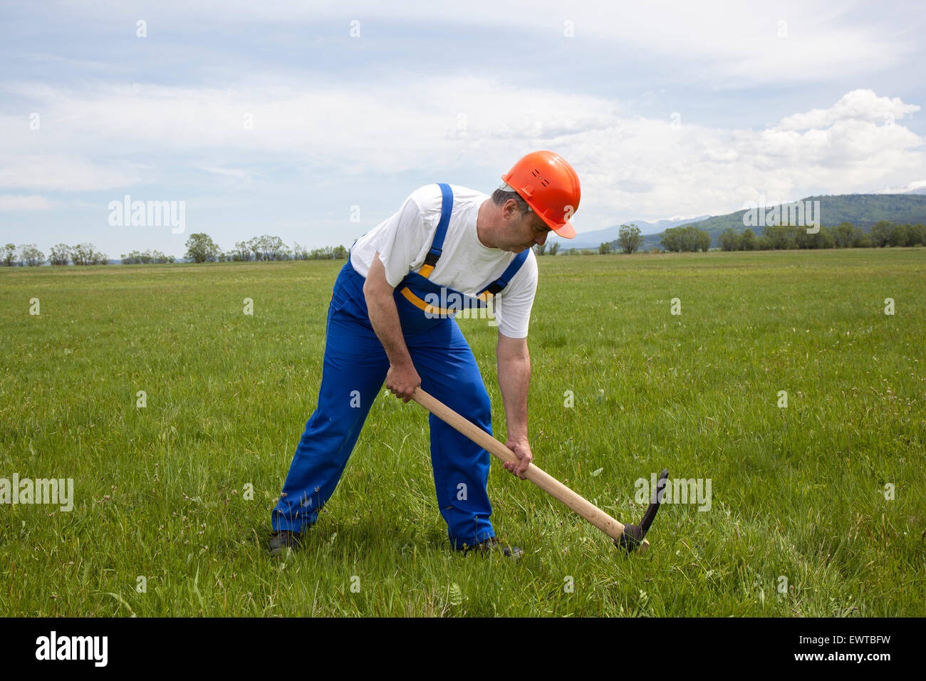Image of a worker in work clothes and hardhat digging with a pickaxe on the green field. Stock Photo