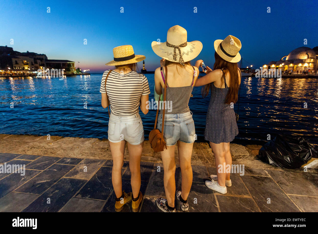 People Three young women in straw hats Chania harbour Crete tourists Greece at dusk Crete Chania Greece Rear View Sunset Scenery Stock Photo