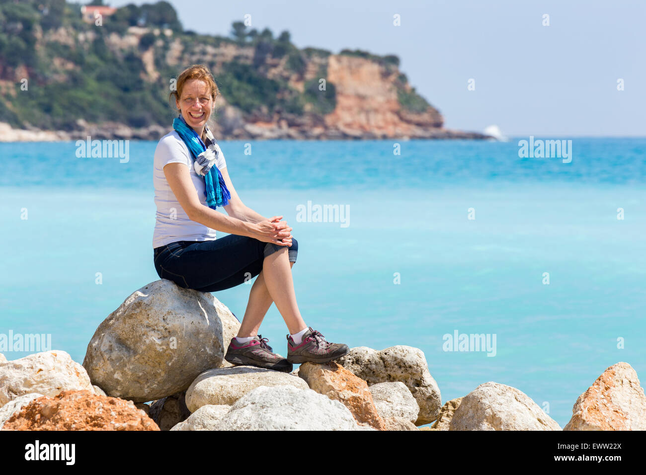 Caucasian middle aged woman as tourist sitting on rocks near blue sea enjoying vacation Stock Photo