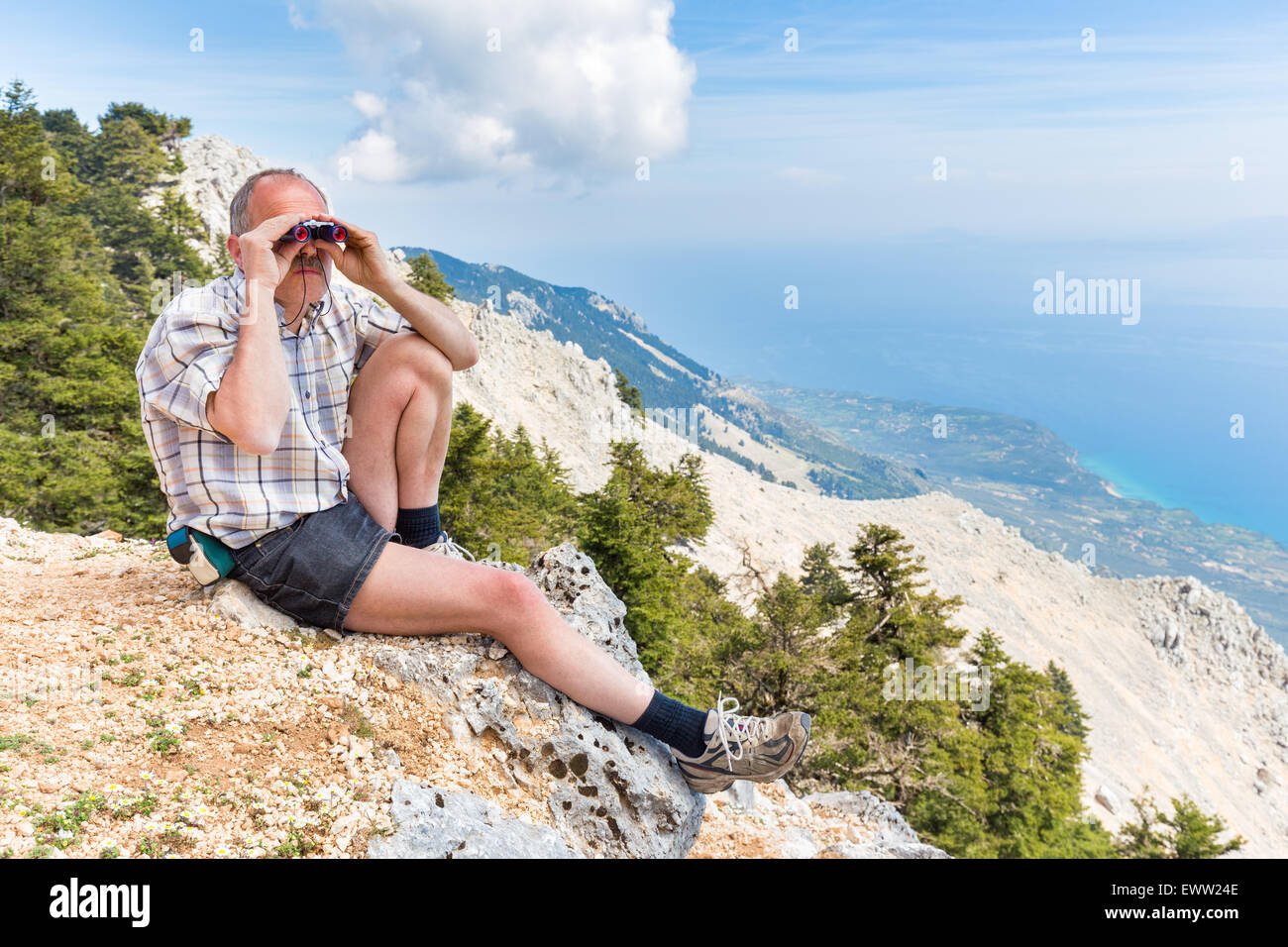 European middle aged man sitting on rock in mountains looking at landscape through binoculars Stock Photo