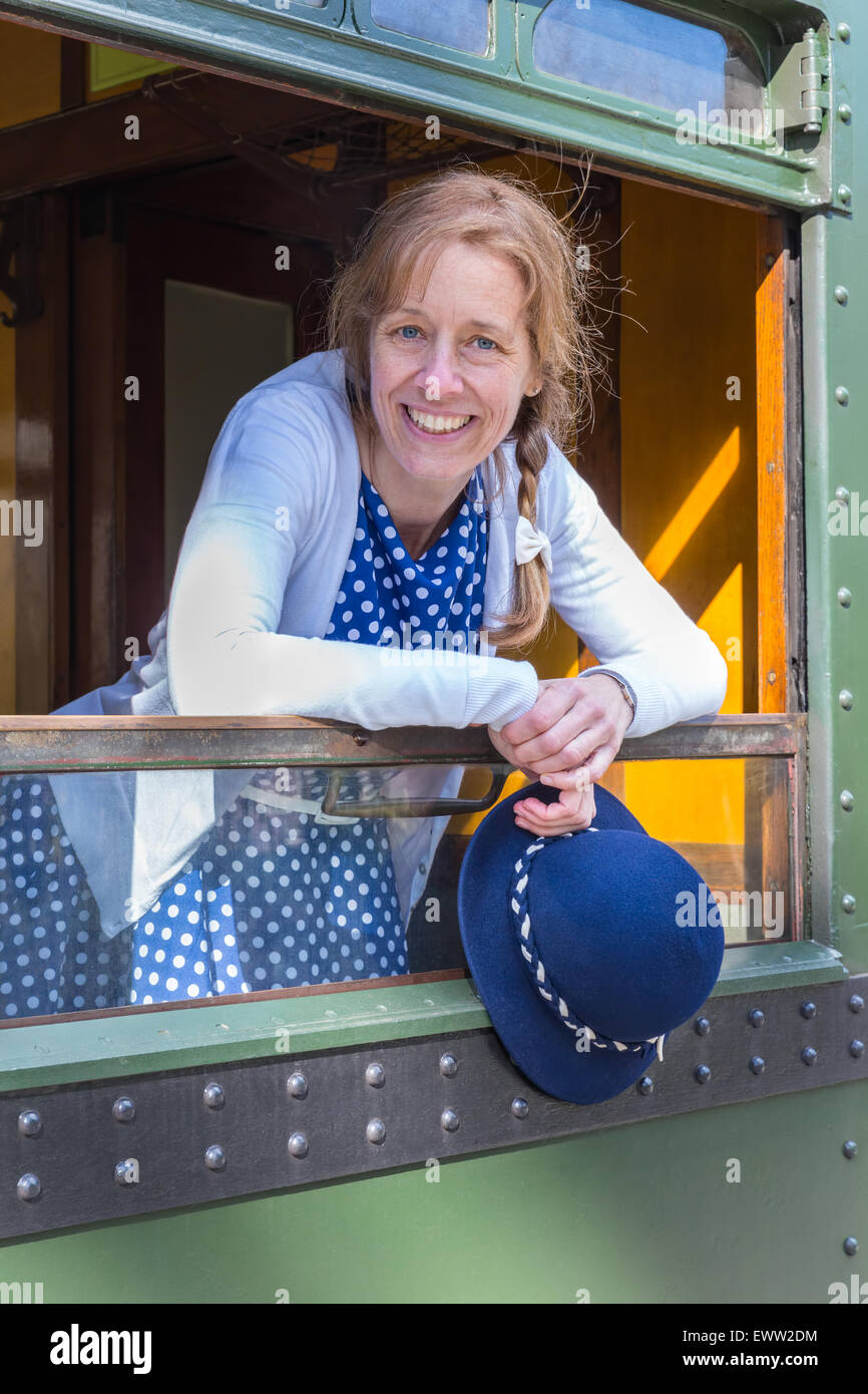 Caucasian middle aged woman dressed in blue old-fashioned clothes holding hat in train window Stock Photo
