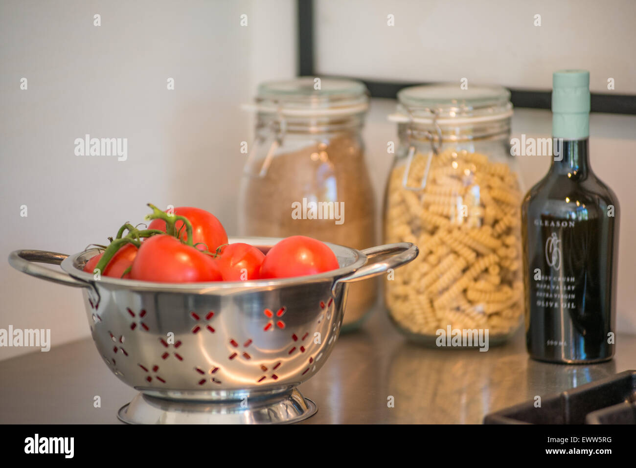 Colander containing ripe tomatoes, a bottle of olive oil, and a jar of rotini on a stainless steel kitchen countertop Stock Photo