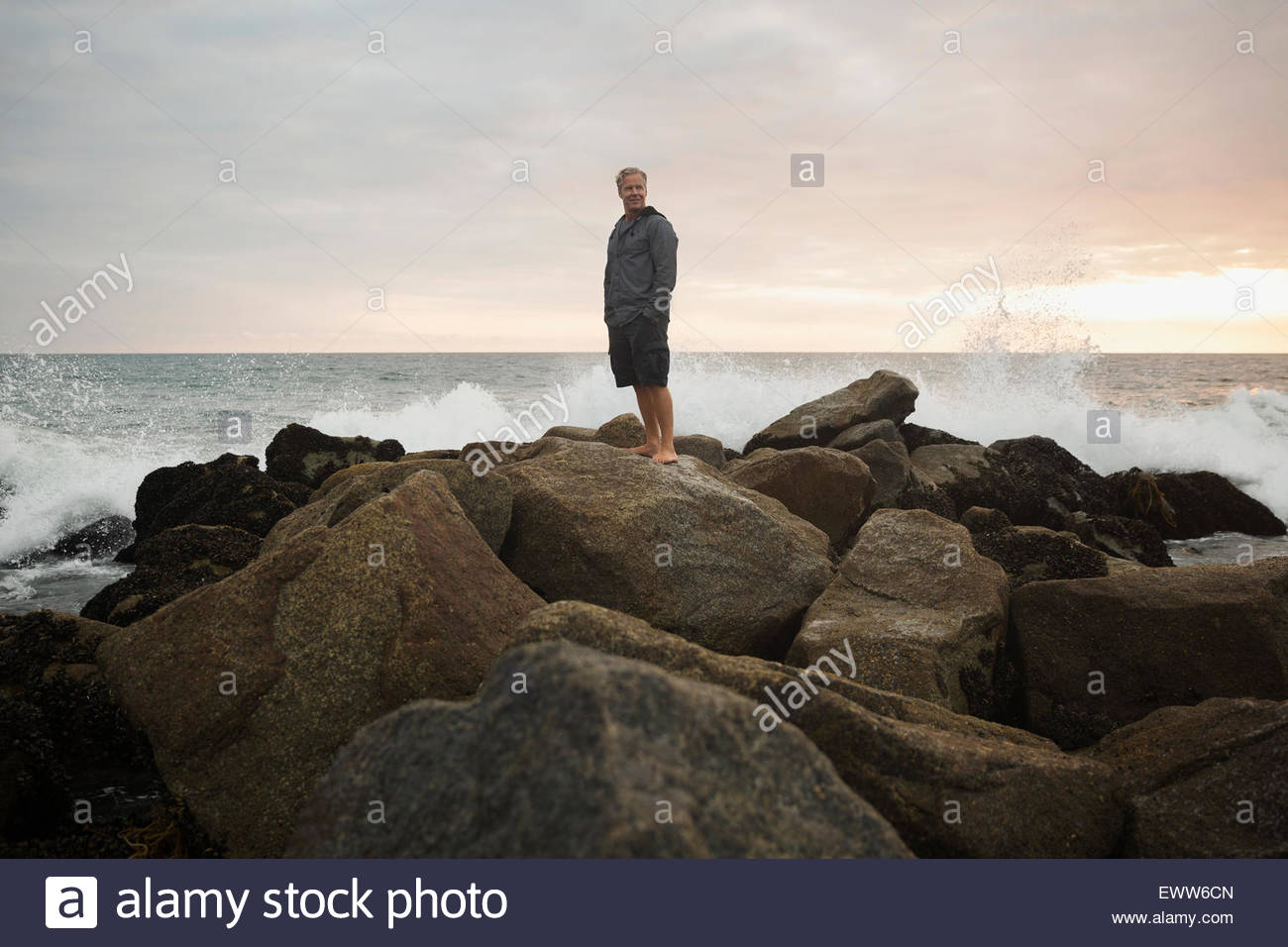 Ocean splashing rocks behind man at sunset Stock Photo