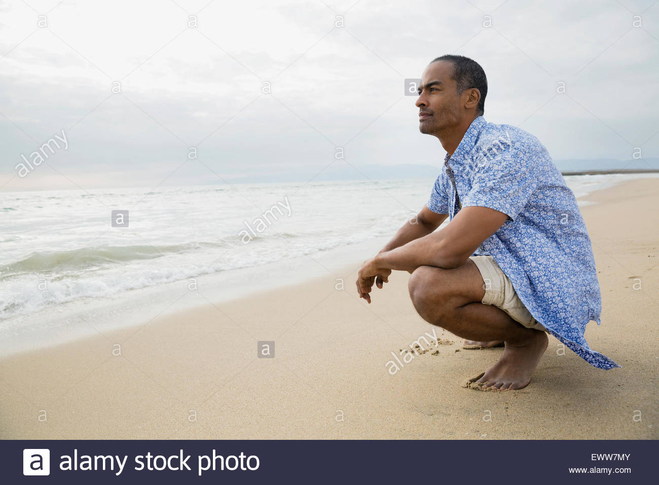Pensive man crouching on beach looking at ocean Stock Photo