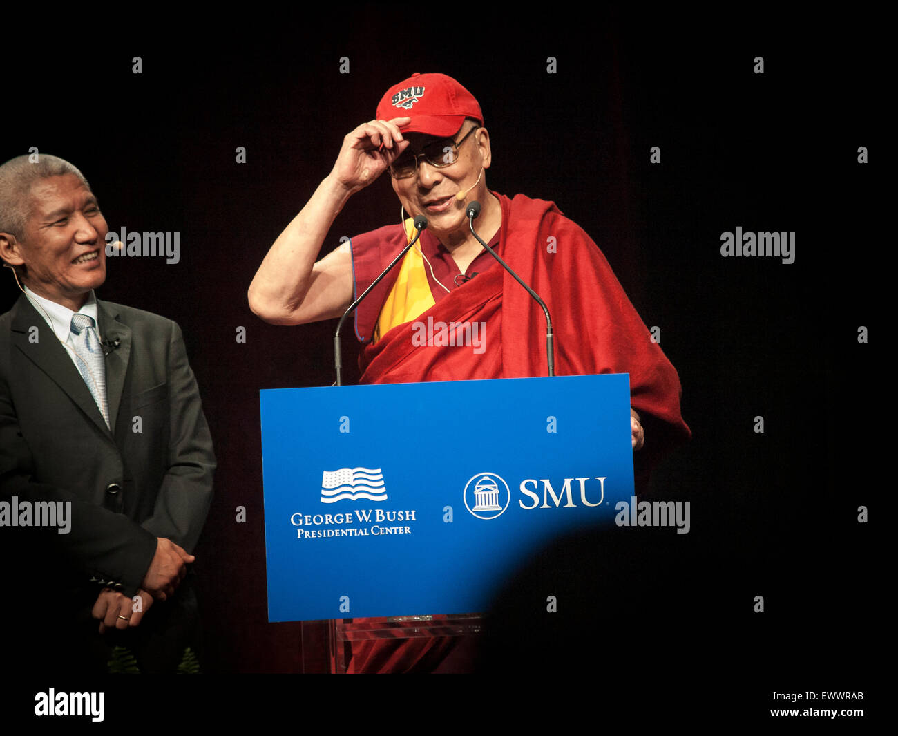 Dallas, Texas, USA. 1st July, 2015. Dalai Lama returns to Southern Methodist University in Dallas, Texas and wears an SMU baseball cap to shield his eyes from bright lights on stage in front of 5200 people. His translator stands next to him. Credit:  J. G. Domke/Alamy Live News Stock Photo