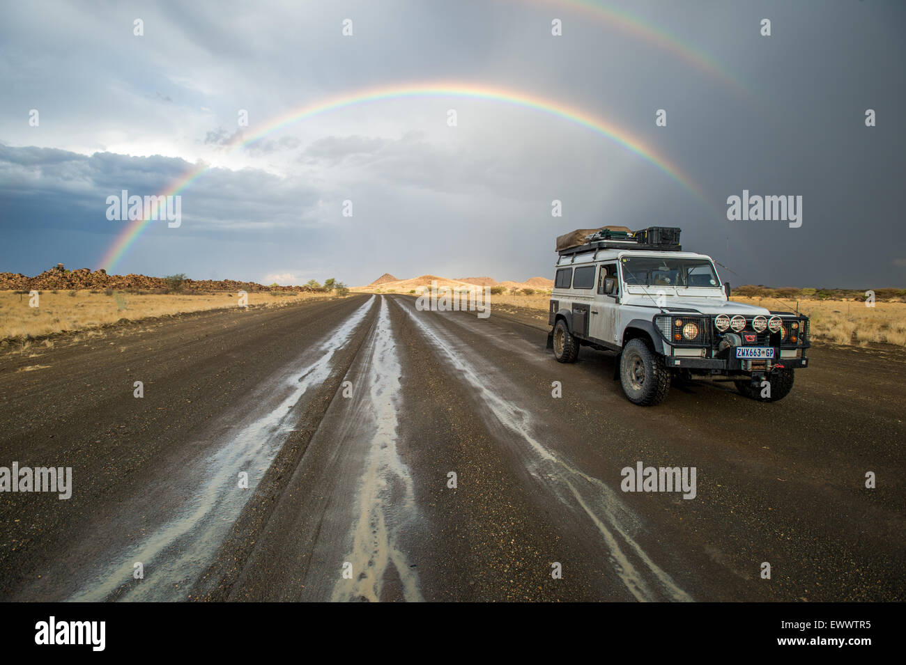Keetmanshoop, Namibia, Africa - Land Rover driving down wet road with rainbow arching in the sky behind it Stock Photo