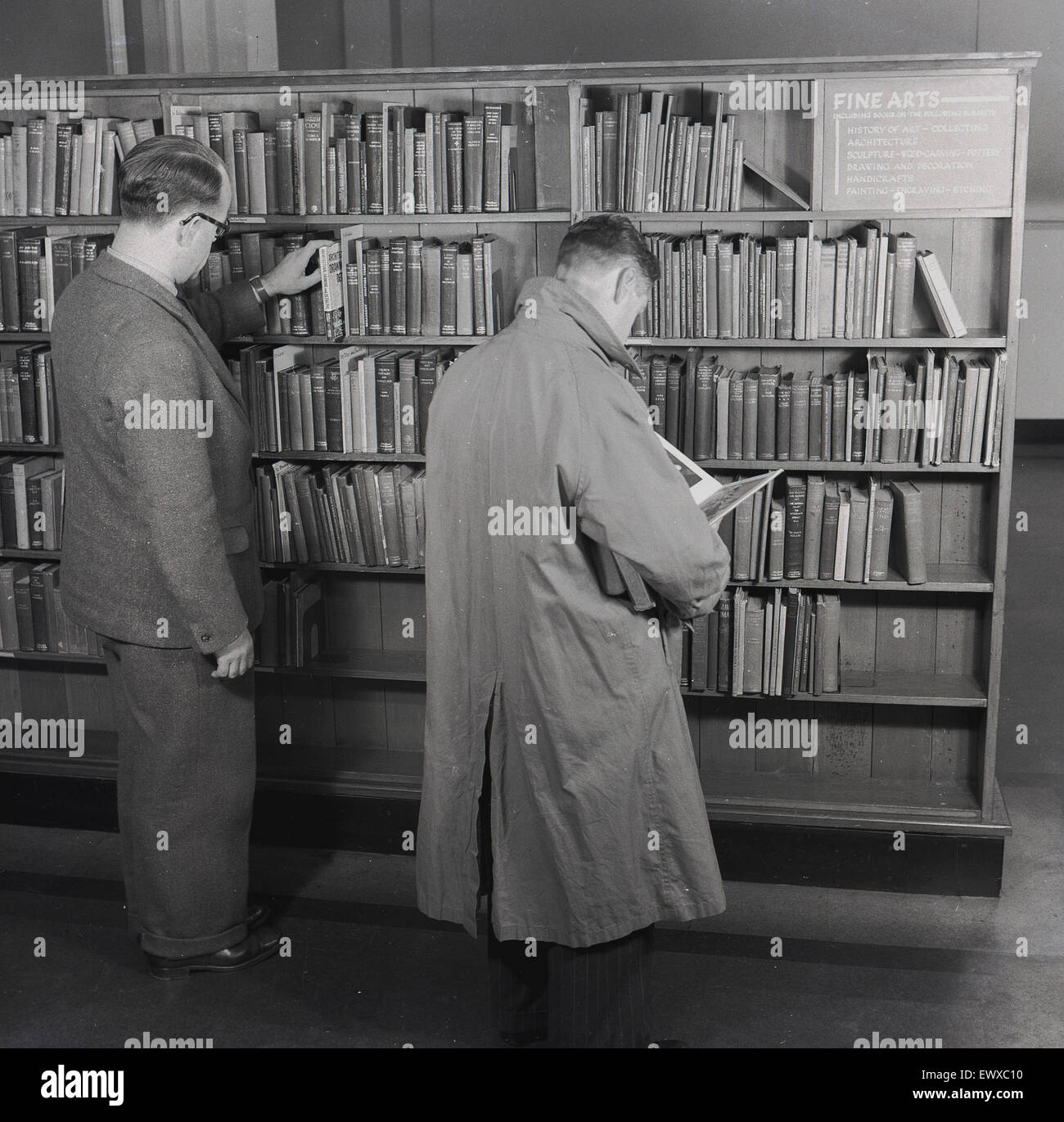 1950s, historical, two men in a public library standing at a bookcase of reference or text books available in the Fine Arts section, one looking and the other, in a raincoat, reading a book, England, UK. Stock Photo