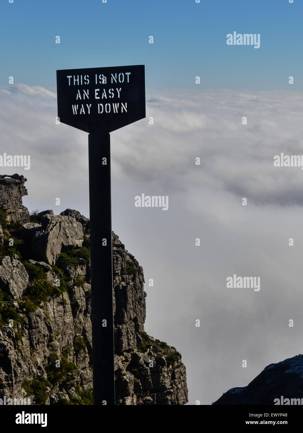Sign reading 'This is not an easy way down' at the edge of Table Mountain, Cape Town, in front of a carpet of white clouds Stock Photo